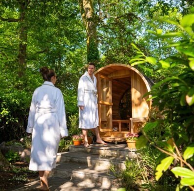 Guests in robes enjoy a serene garden sauna under lush trees, highlighting relaxation and nature-focused amenities.