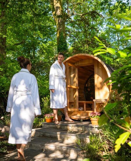 Guests in robes enjoy a serene garden sauna under lush trees, highlighting relaxation and nature-focused amenities.