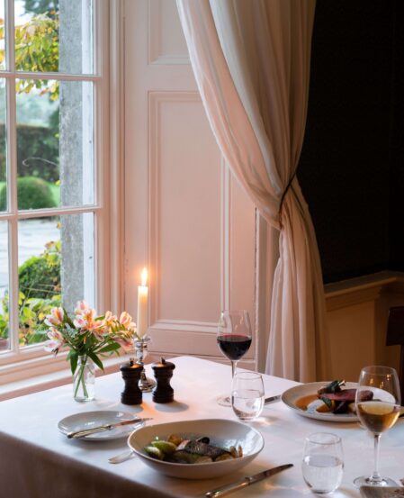 Cosy hotel dining with candlelit table, elegant curtains, garden view; features wine, gourmet dishes, perfect for romantic dinners.