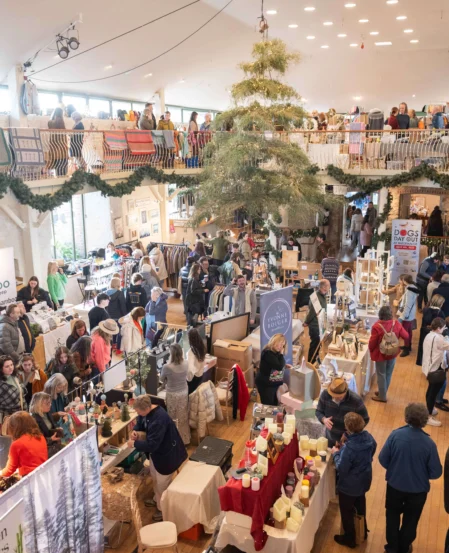 Festive indoor market in a hotel atrium with stalls, balcony seating, central Christmas tree and guests browsing local crafts and food.