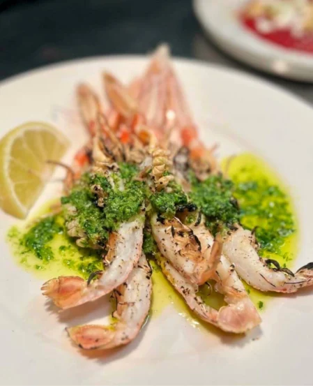 Grilled langoustines on white plate with green herb sauce, olive oil drizzle and lemon wedge — seaside restaurant dining amenity